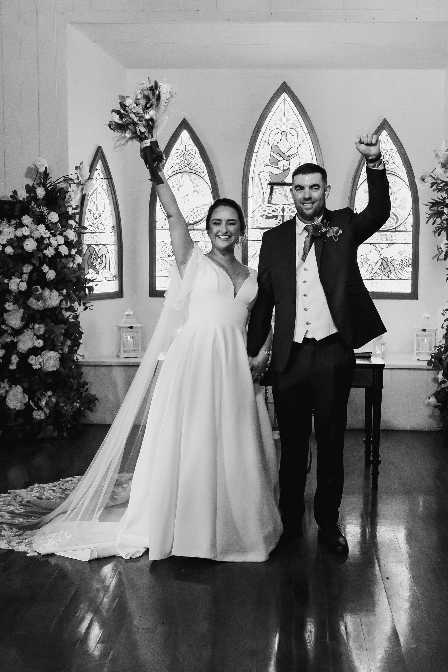Joyful bride and groom celebrate just married moment raising hands in chapel with gothic windows.