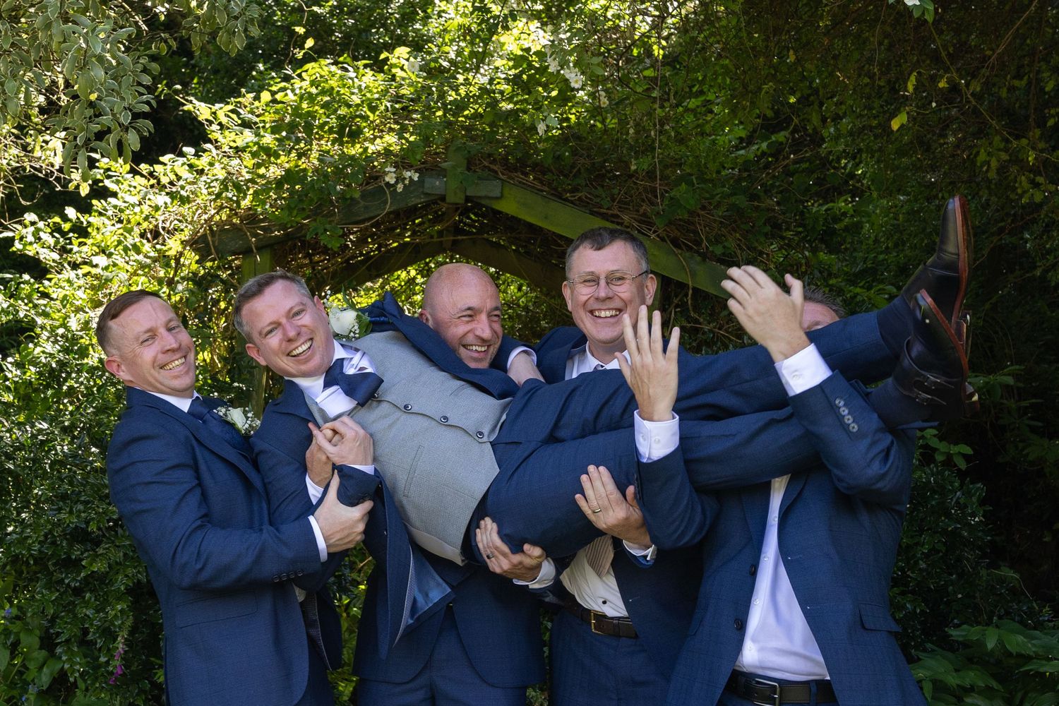 Four groomsmen in navy suits joyfully lifting the groom outdoors at a wedding celebration.