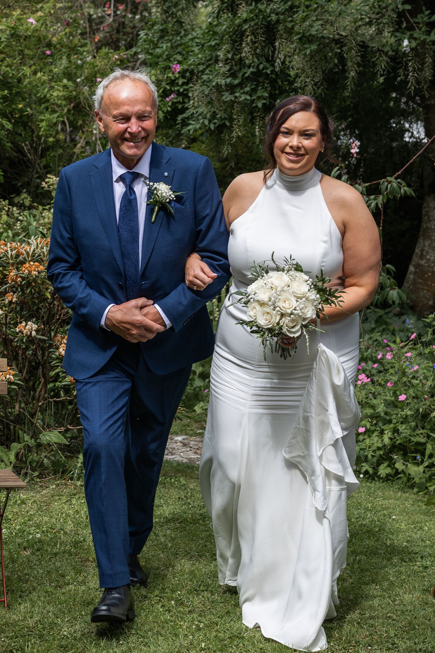 Bride in white gown holding bouquet walks with older man in blue suit through garden on wedding day.