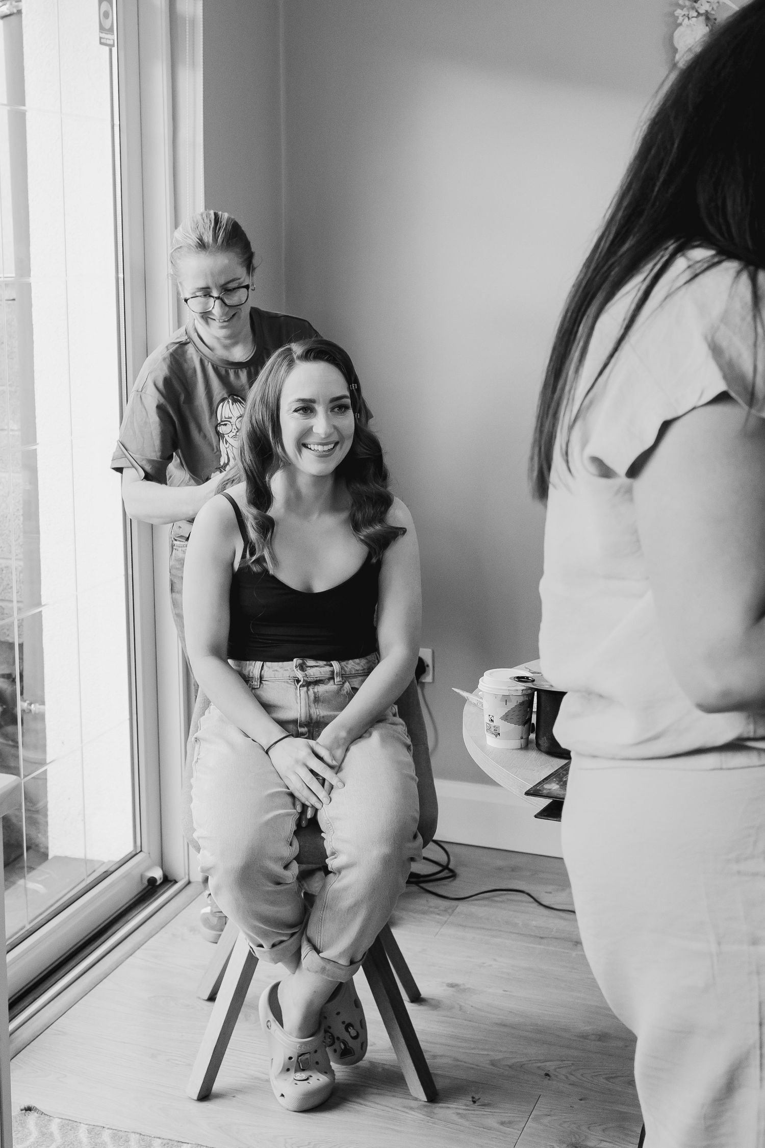 Black and white photo of someone sitting on a chair in casual attire by a window.