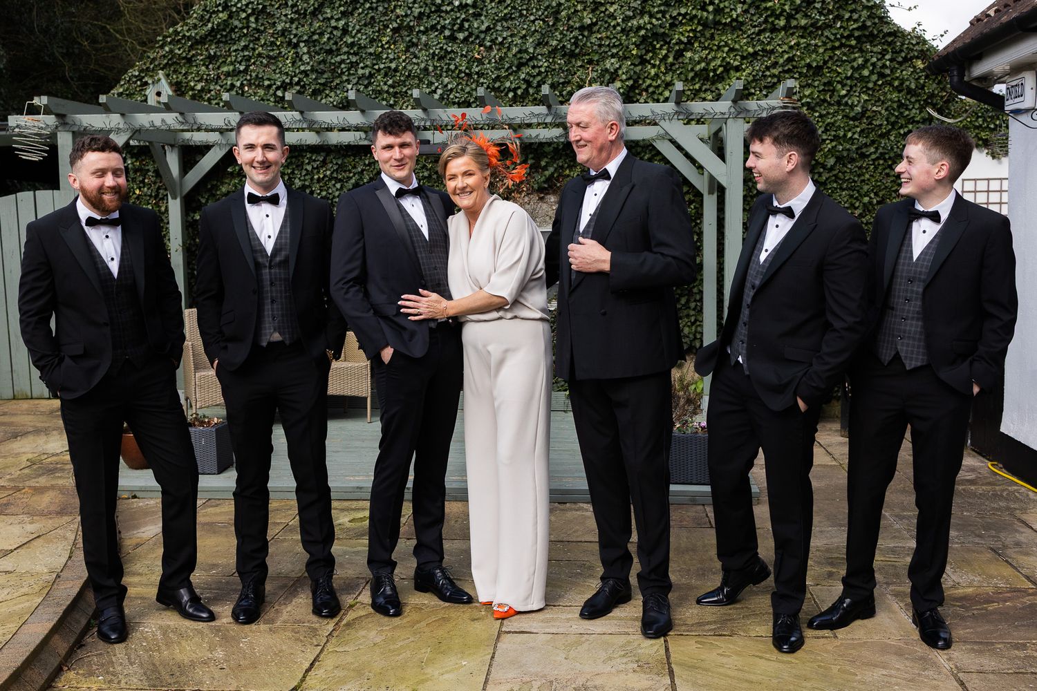 Wedding party in black tuxedos and formal attire posing against Dublin garden wall.