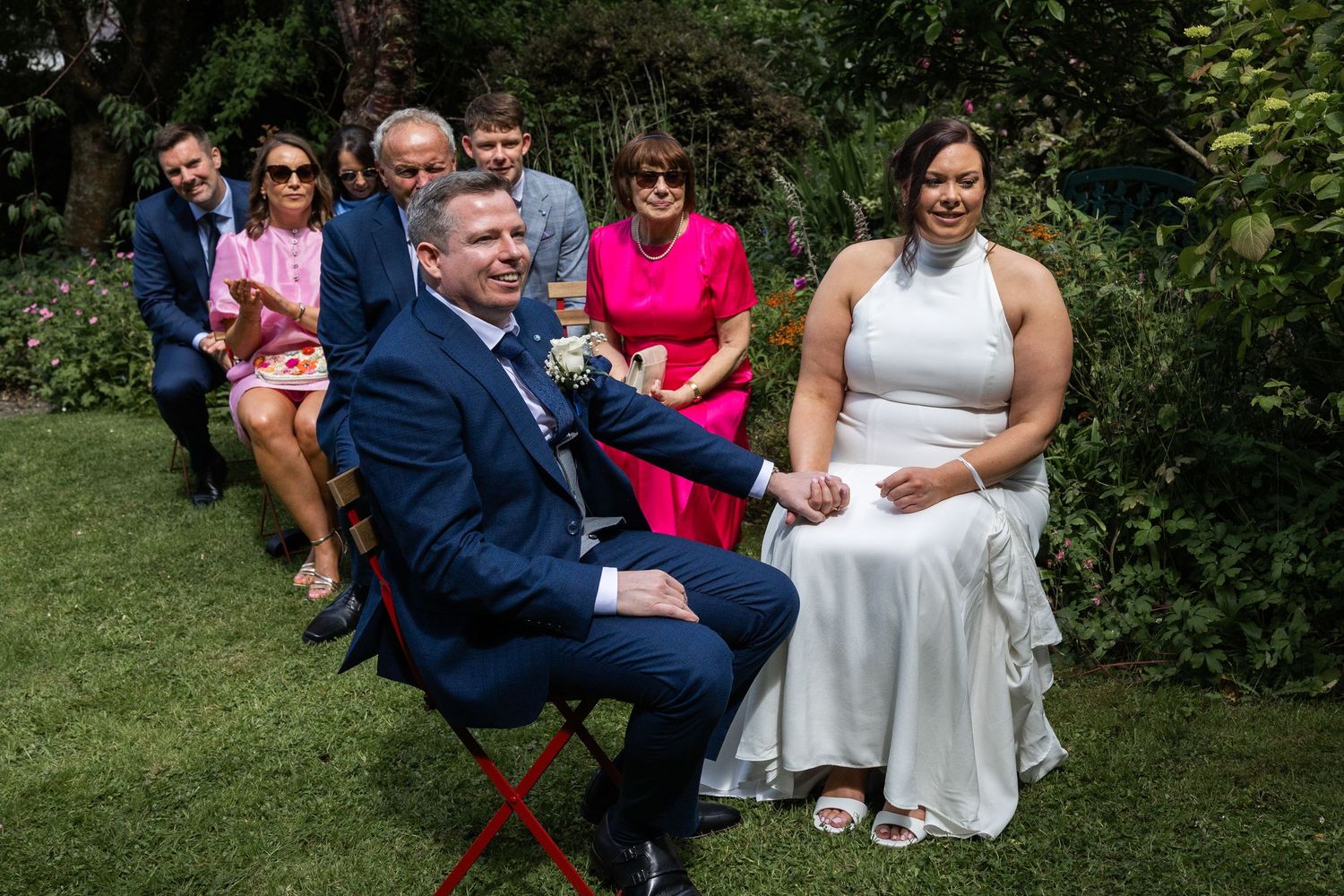 Bride and groom seated outdoors during garden wedding ceremony with guests in background.