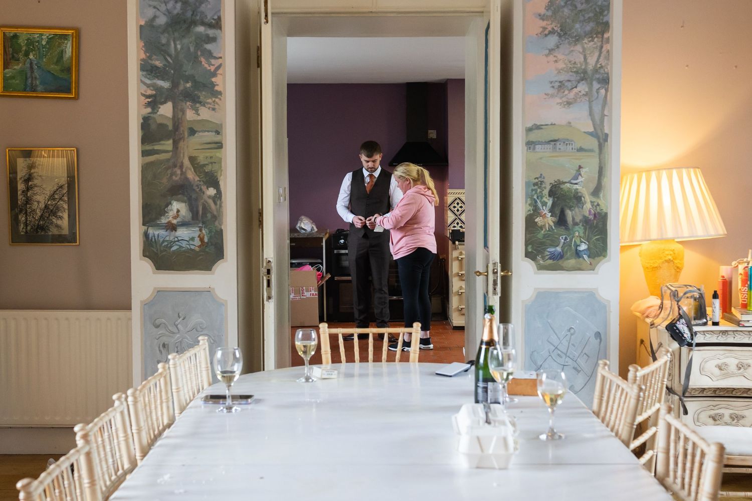 Elegant dining room with long white tablecloth, chiavari chairs, and staff preparing in doorway with scenic wall murals.