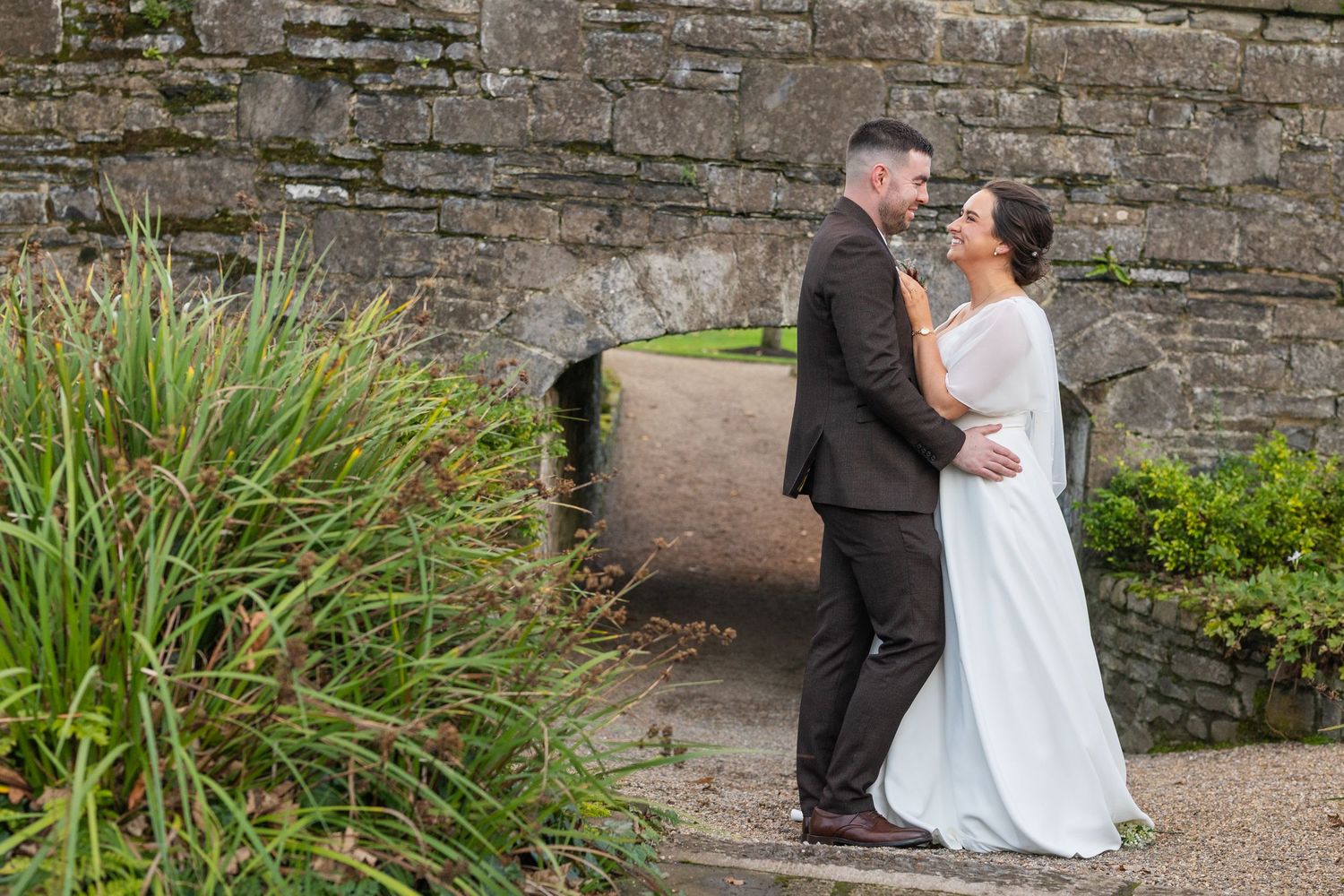 Bride and groom embracing near a stone bridge in a lush garden on their wedding day.