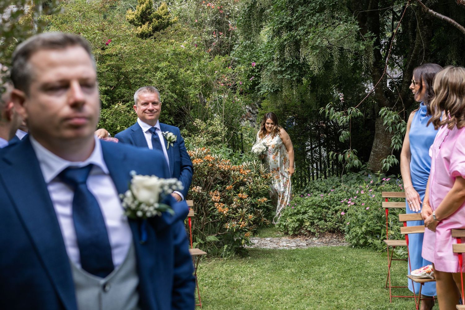 Outdoor garden wedding ceremony with groom in navy suit awaiting bride walking down the aisle.