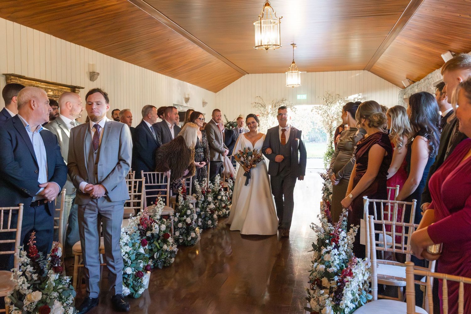 Bride walks down the aisle with escort past floral arrangements at elegant indoor wedding ceremony.