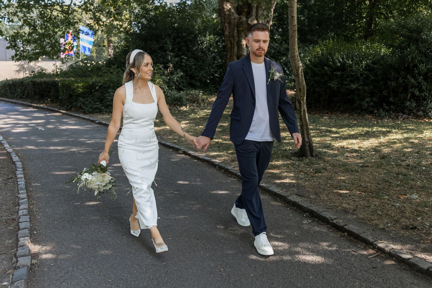 A bride and groom walk hand in hand along a tree-lined path on their wedding day.