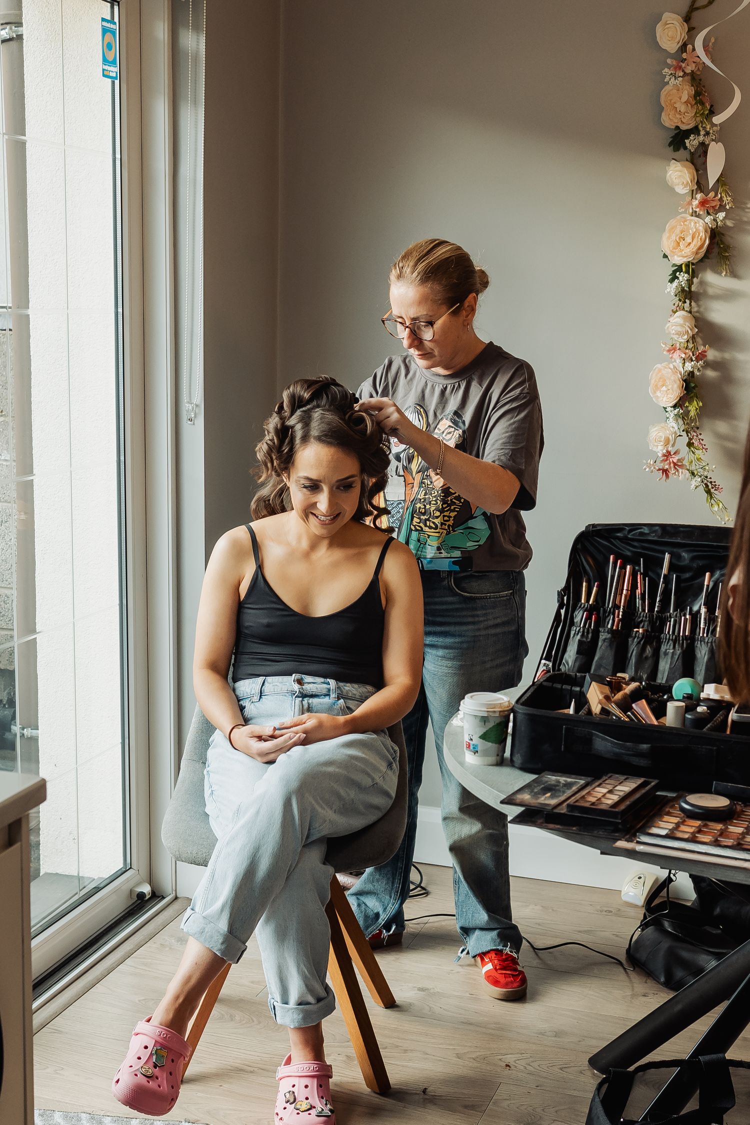 A hairstylist works on a smiling bride-to-be seated near a window, with makeup tools displayed nearby.