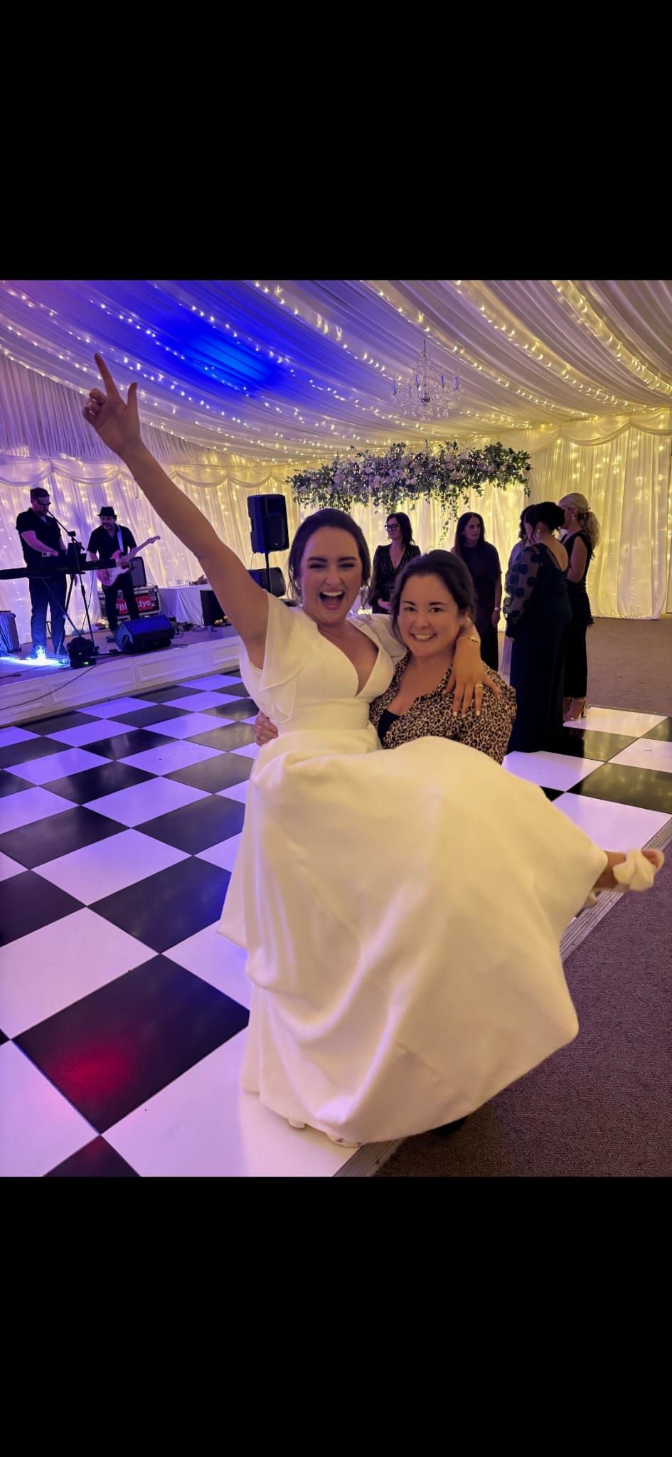 Two people celebrating on a black and white checkered dance floor under twinkling lights at an indoor wedding reception.