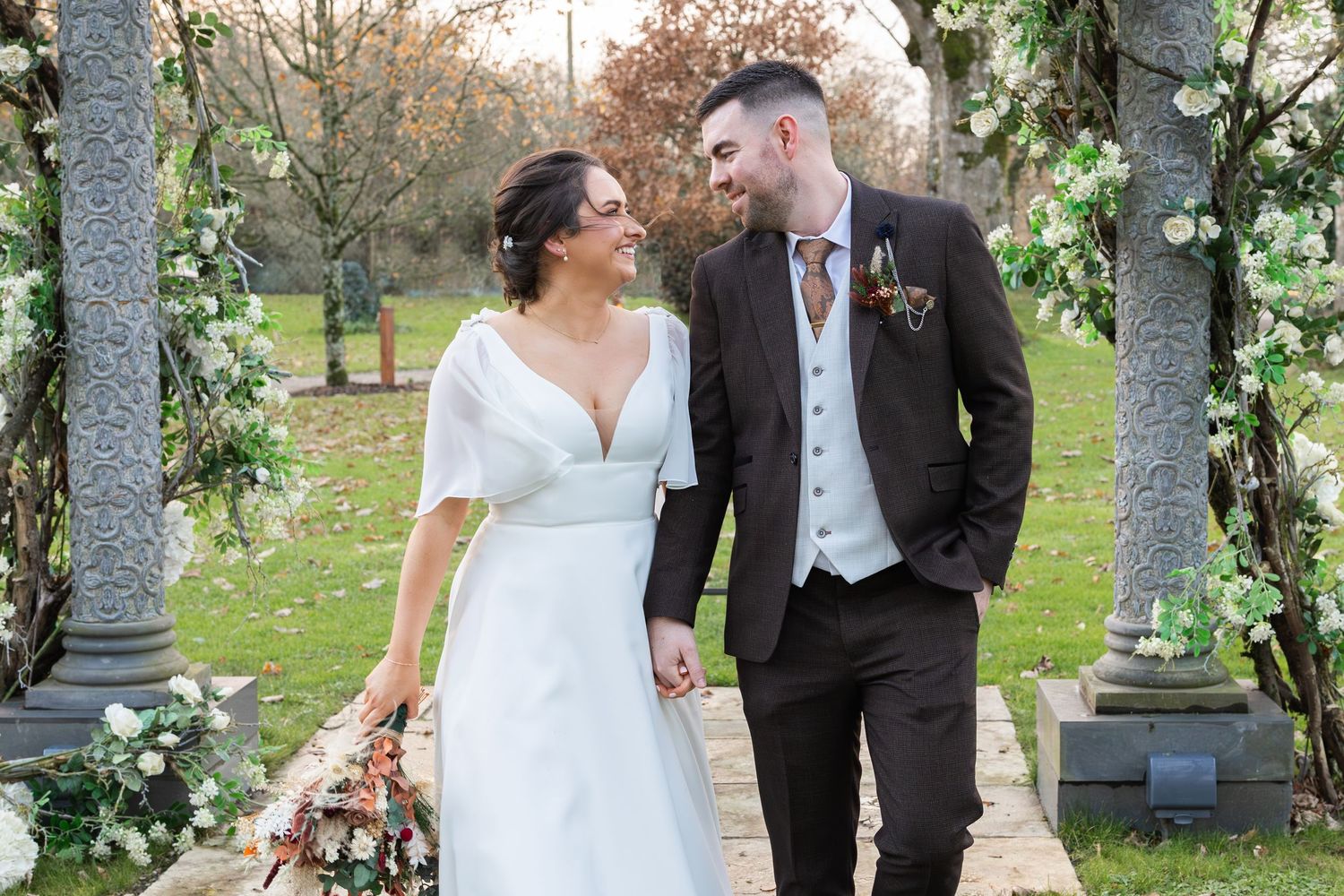 Happy bride and groom walk hand in hand under floral arch in outdoor garden wedding setting.