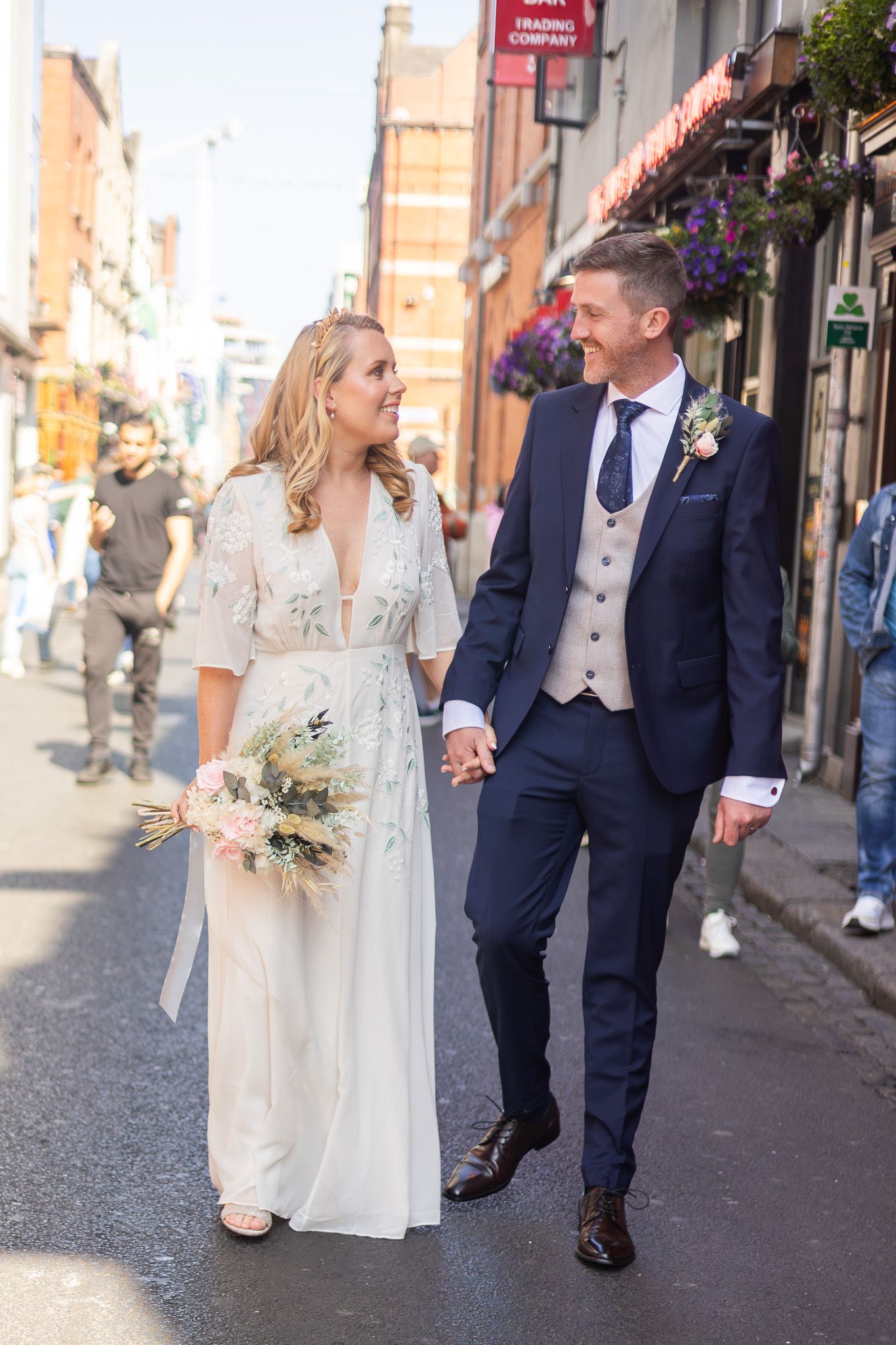 A couple strolls down a charming street with hanging flower baskets on their wedding day.