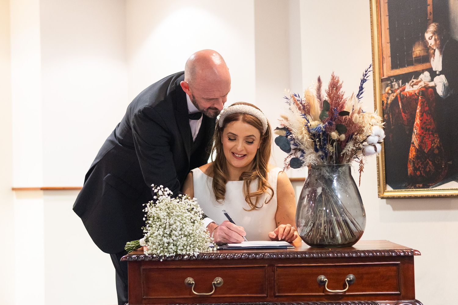 Wedding party members sign documents at an elegant wooden desk with floral arrangements.