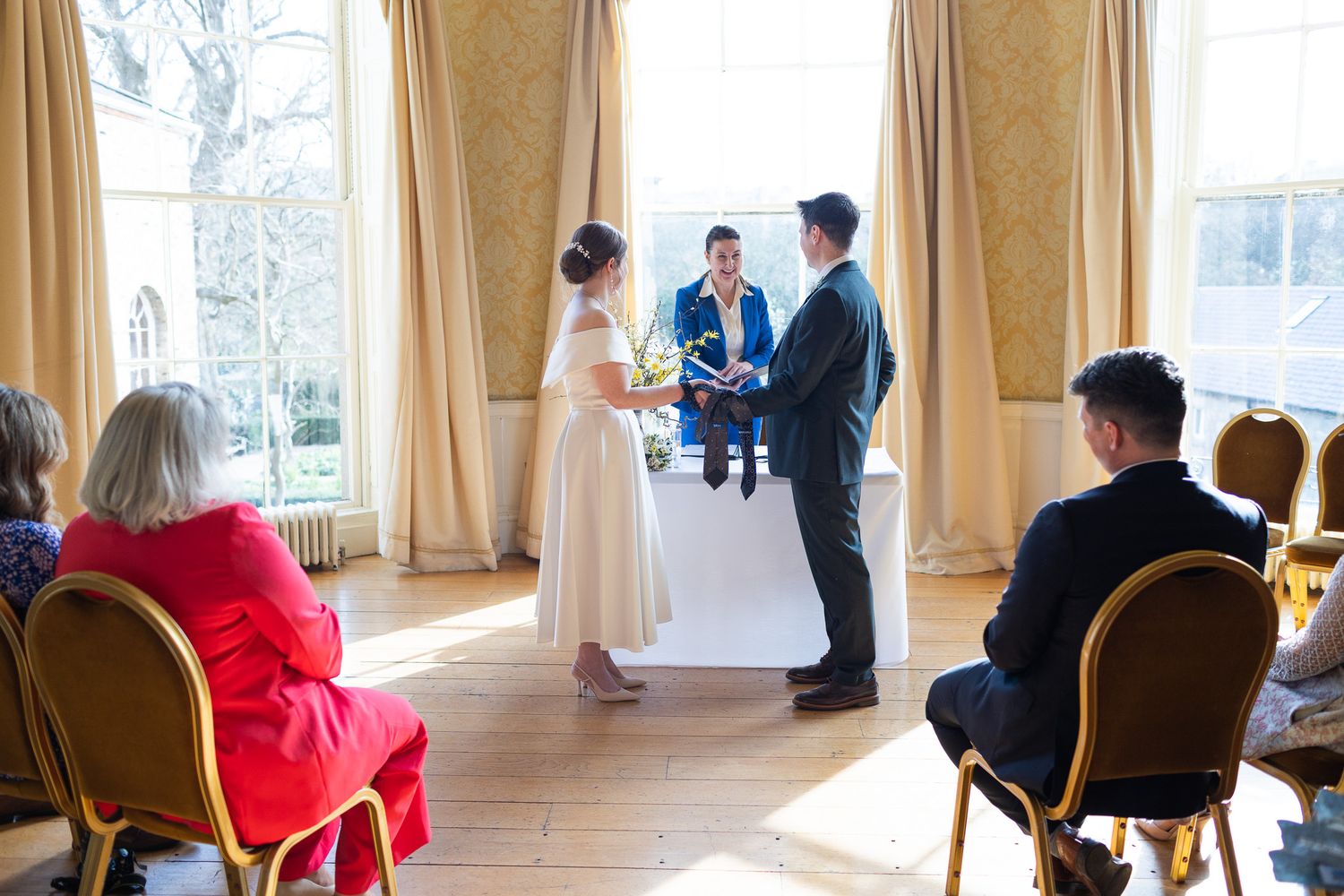 Bright sunlit MoLI  Ceremony room with floor-length curtains and guests seated in gold chairs.