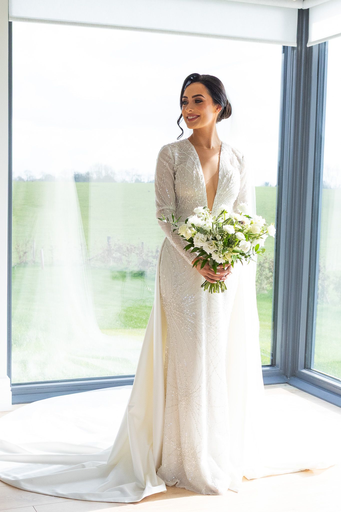 Bride in a white lace wedding gown posing near large windows in a bright, modern Dublin wedding venue.