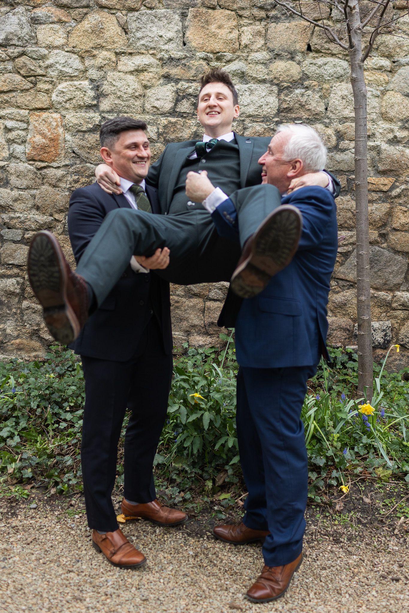 Two people playfully kick their legs up while posing against a stone wall outdoors During relaxed wedding photography in Dublin.