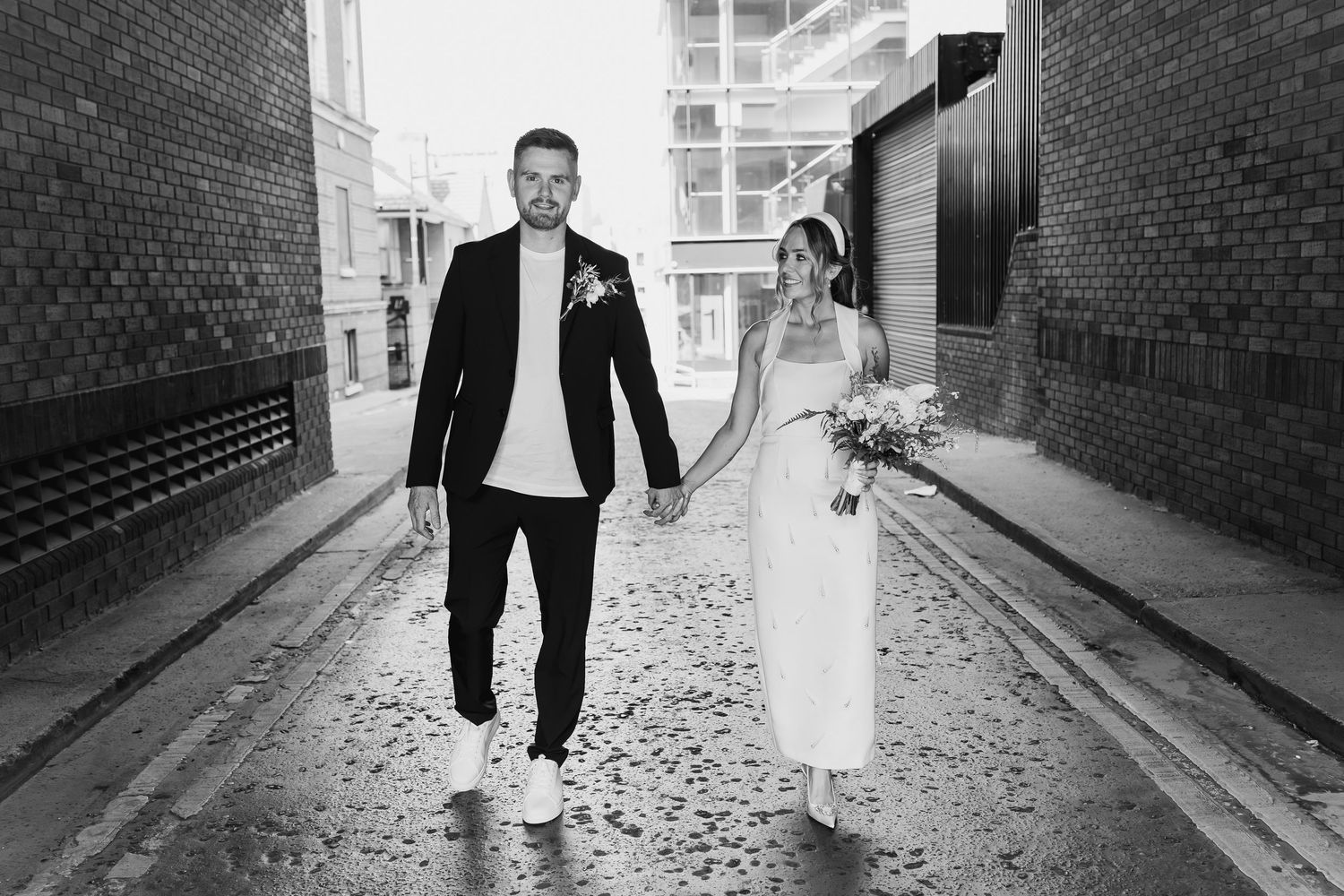 Black and white photo of a bride and groom holding hands walking down an urban alley on their wedding day.