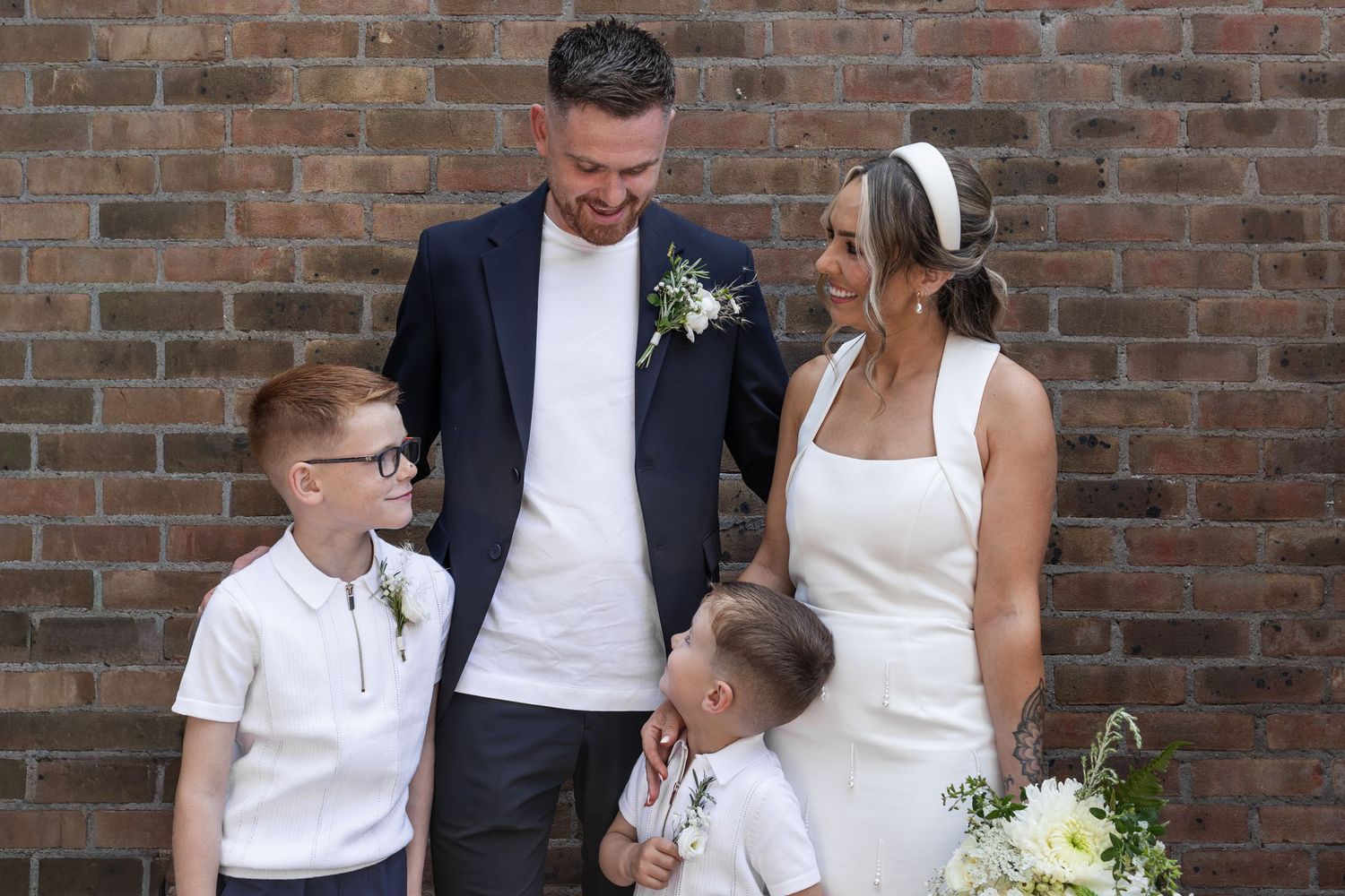 A joyful wedding couple in white and navy stand with two young boys in front of a brick wall.