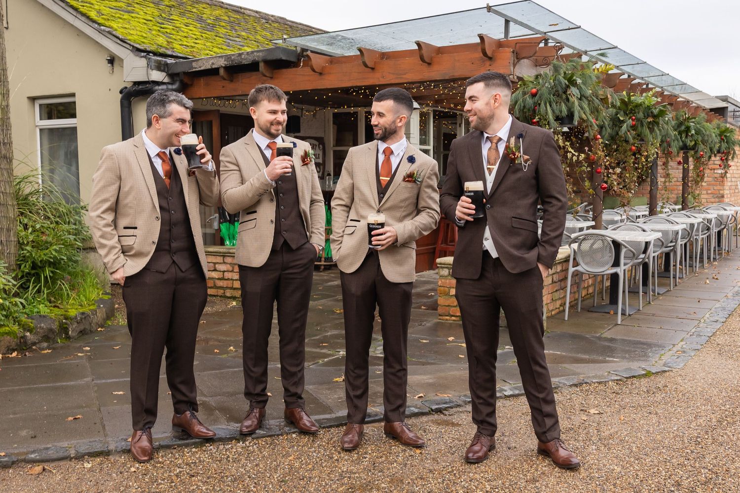 Four groomsmen in brown suits toasting with drinks outside a rustic wedding venue.