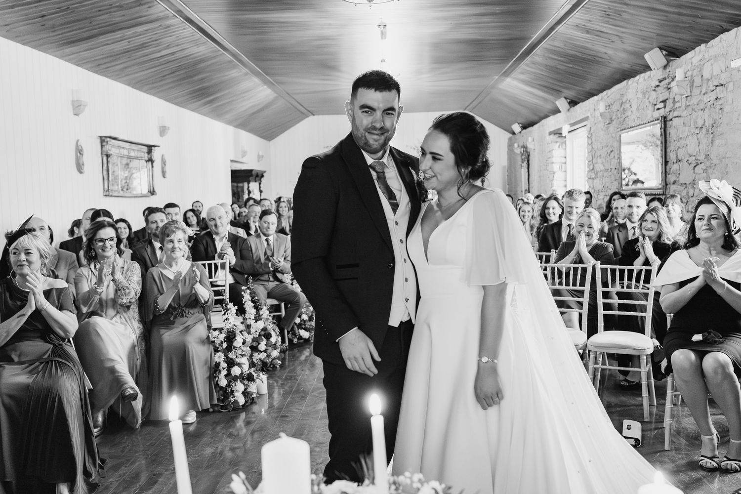 Bride and groom smiling joyfully at wedding ceremony altar, guests applauding in elegant stone venue.