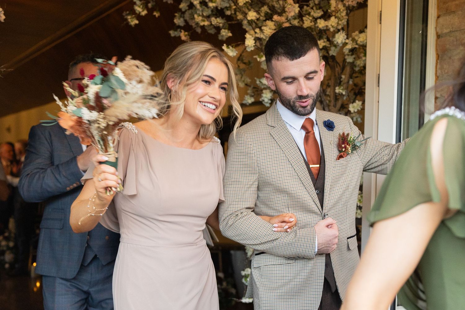 Bride and groom walk arm in arm, smiling, as guests toss dried flowers during wedding ceremony exit.