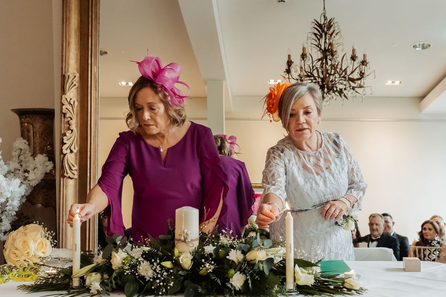 Two elegantly dressed women lighting candles at a floral wedding reception table centerpiece.
