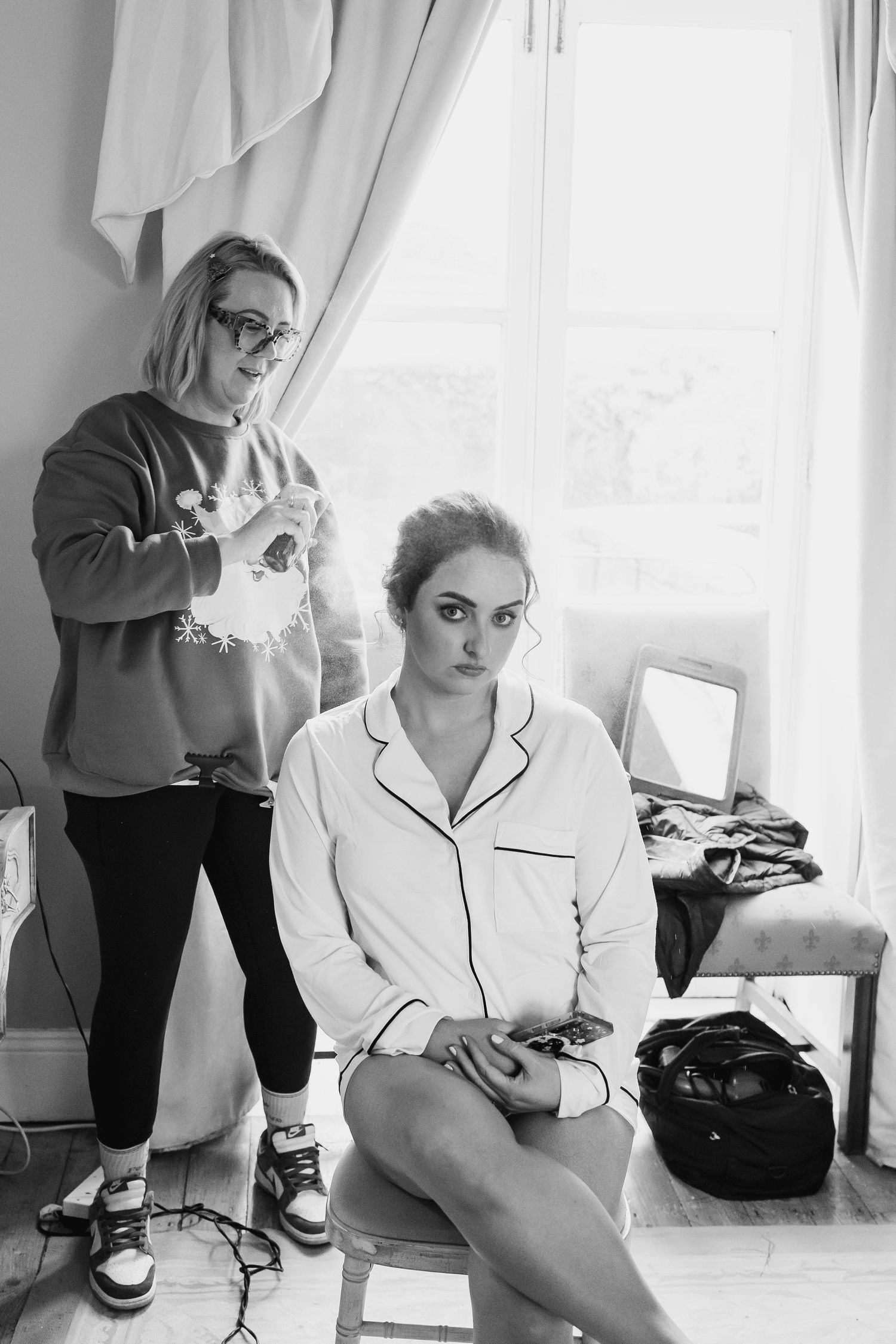Black and white photo of a woman in pajamas sitting while a hairstylist works on her hair in a bright room.