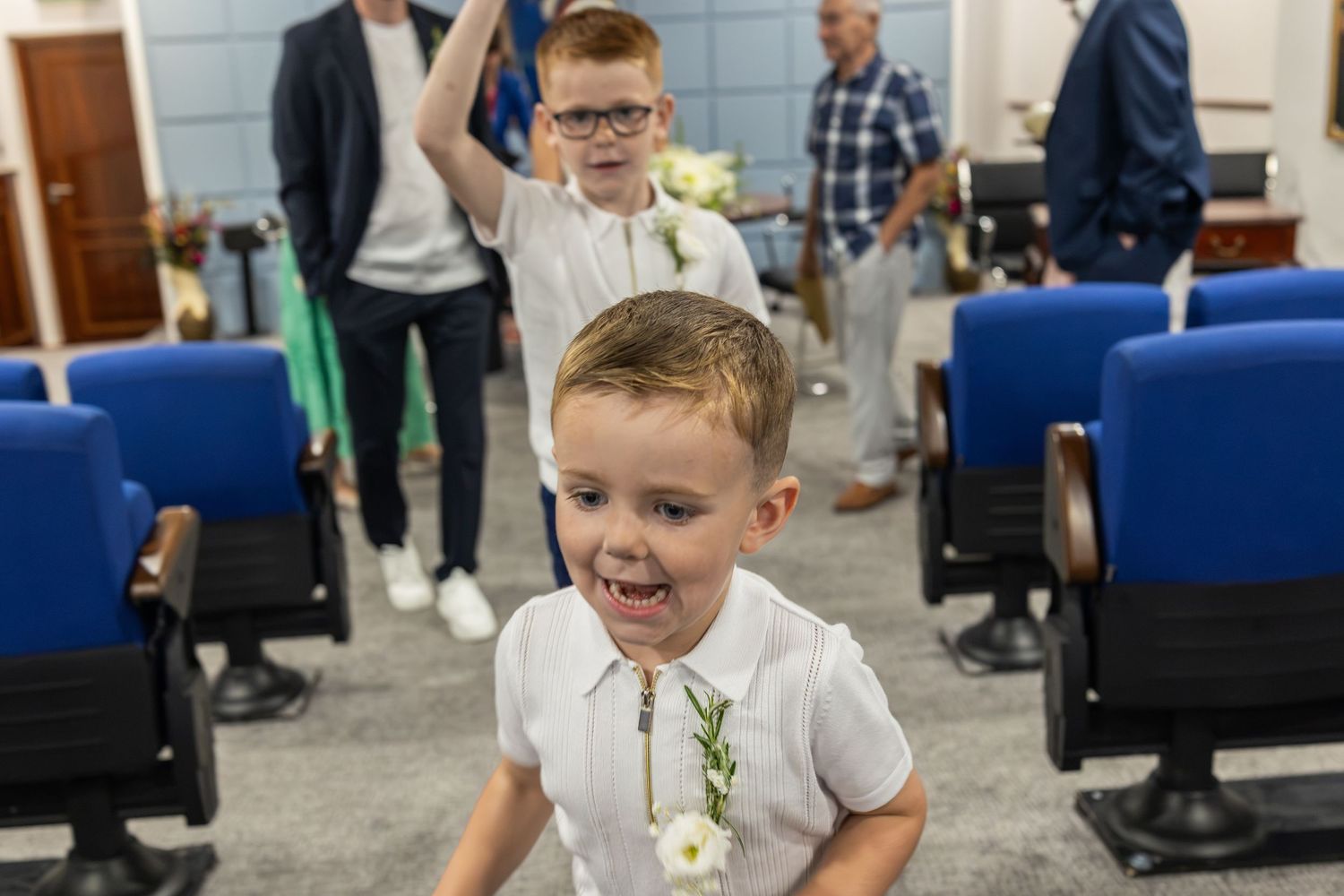 Two young boys in white shirts laugh and play in a wedding venue aisle with blue chairs.