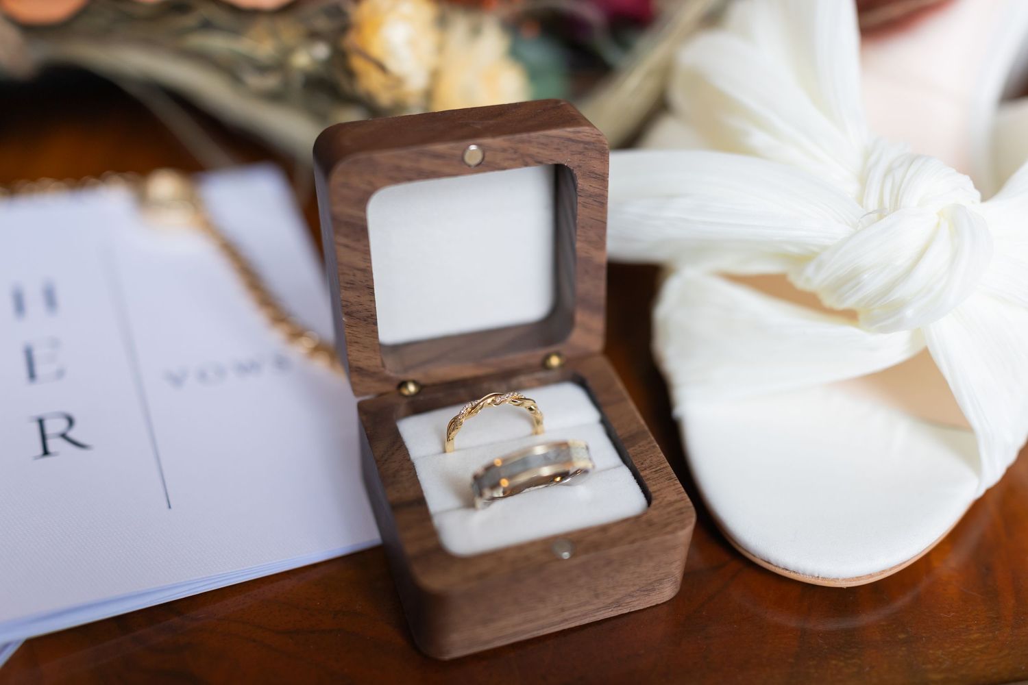 Two gold wedding bands displayed in an open wooden ring box beside a white bridal shoe and wedding stationery.