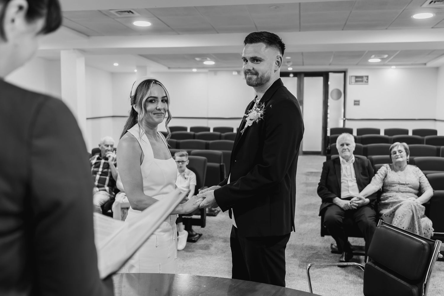 Black and white photo of a couple exchanging vows at a civil wedding ceremony, holding hands before an officiant.
