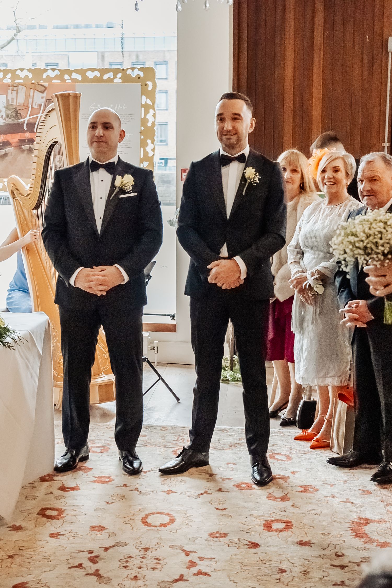 Two grooms in black tuxedos stand at the altar smiling during their wedding ceremony with guests behind them.
