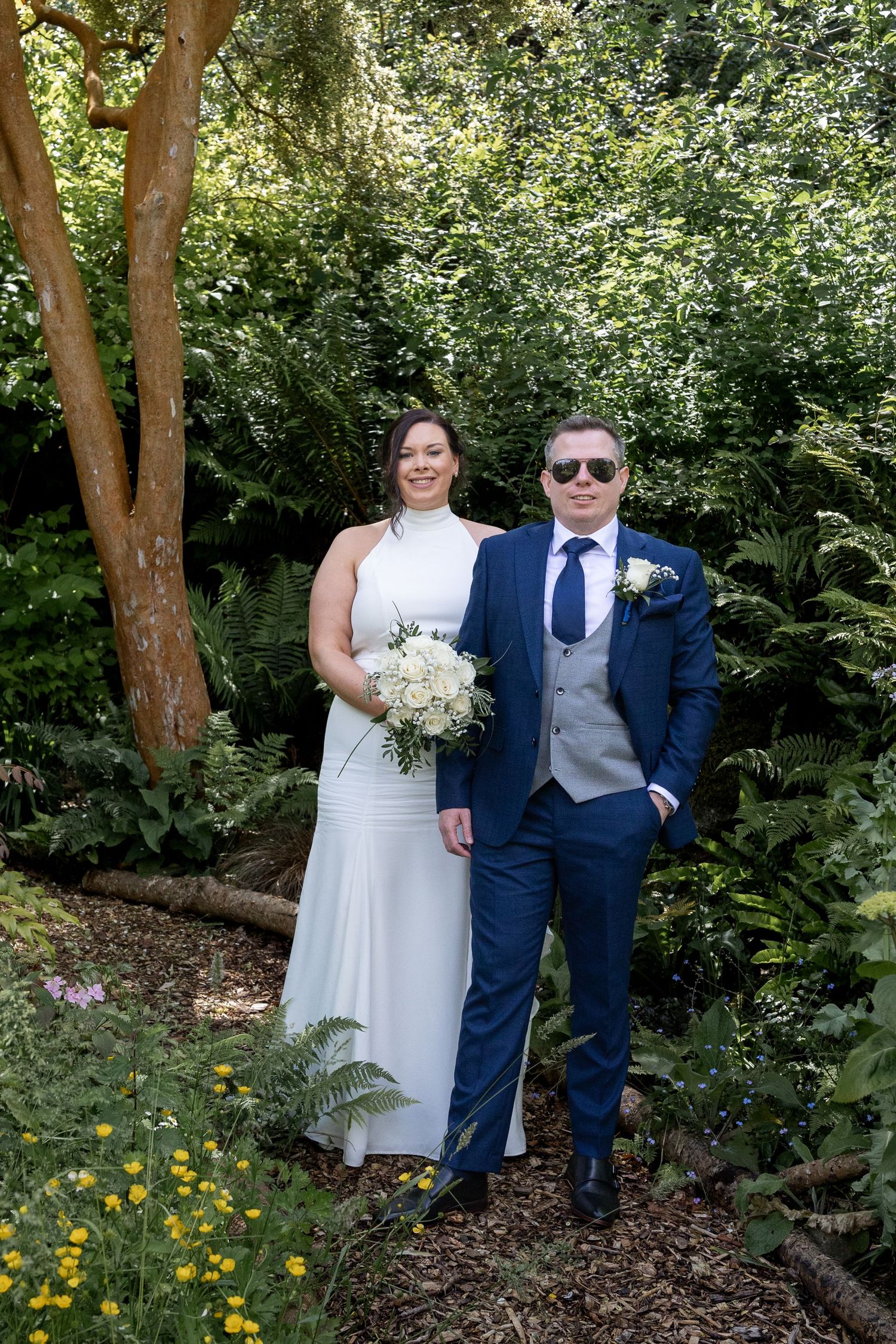 Bride in white halter gown and groom in navy suit with boutonniere pose together in a lush garden setting.