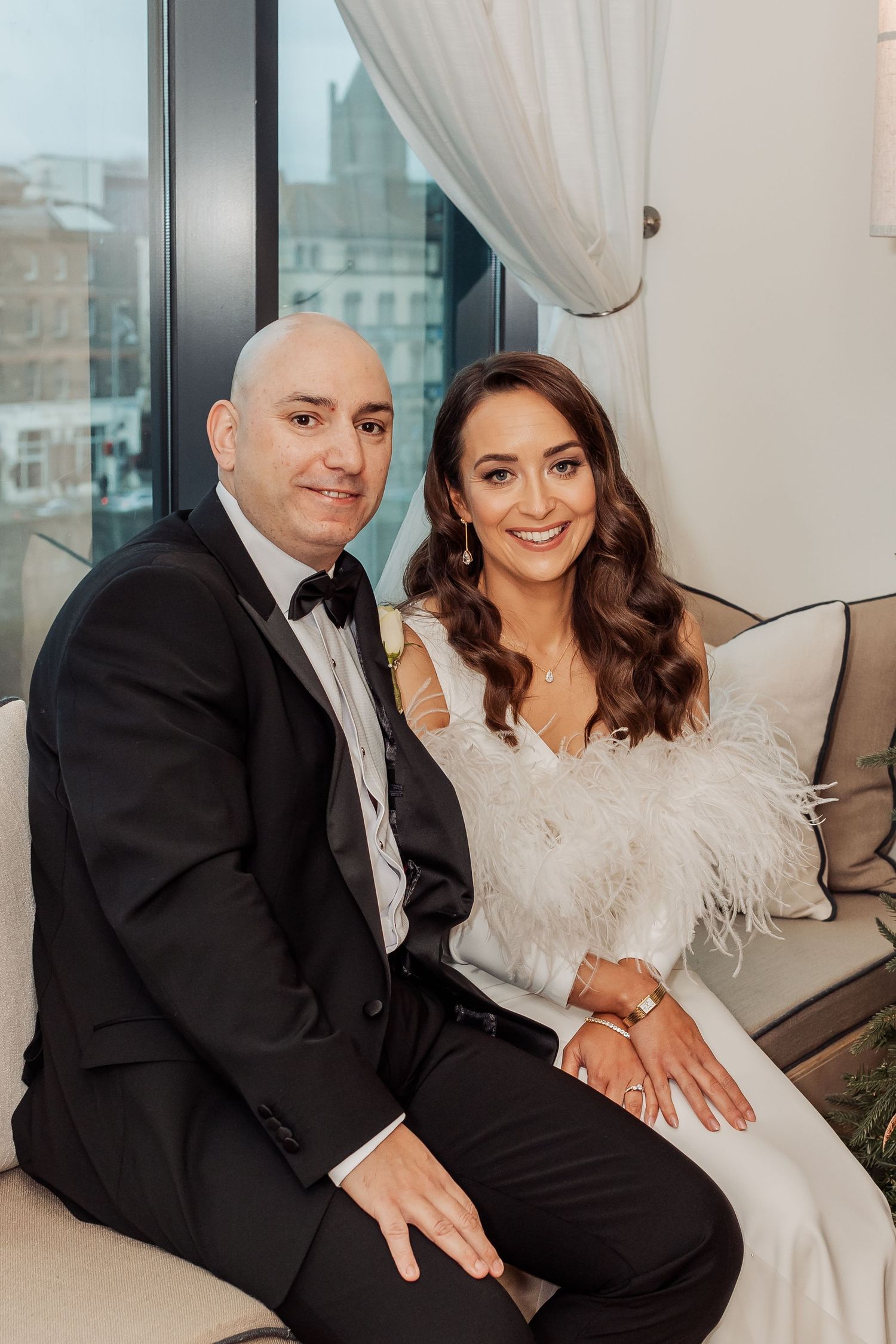 Elegant couple poses together on a couch in formal wedding attire near a window.