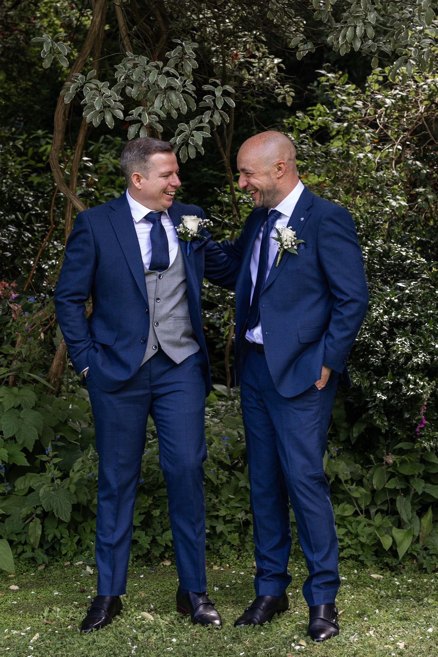 Two grooms in matching navy blue suits with boutonnieres laughing together outdoors on their wedding day.