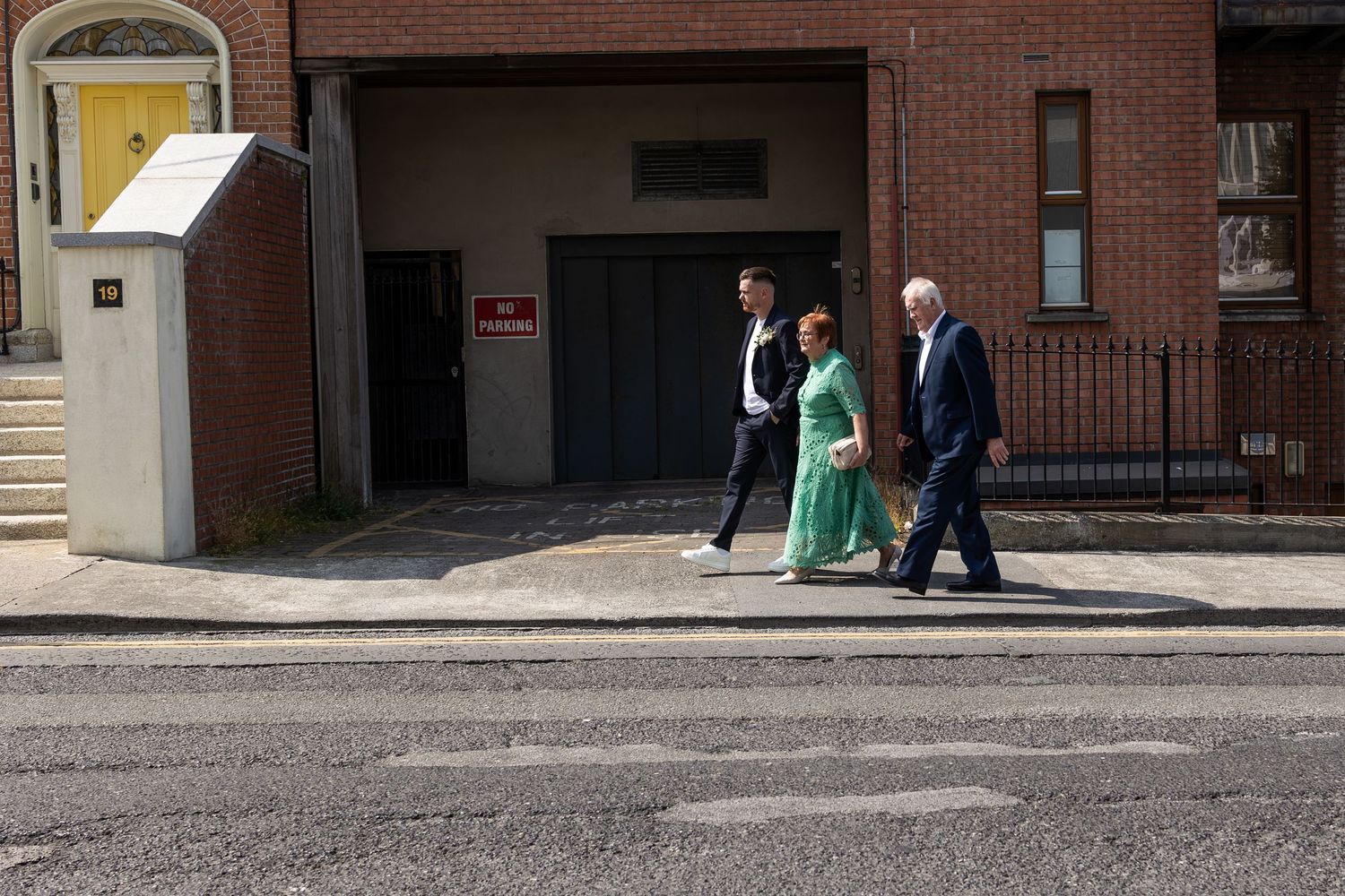 Three people walking past a brick building entrance on a sunny day, woman in green dress flanked by two men in suits.