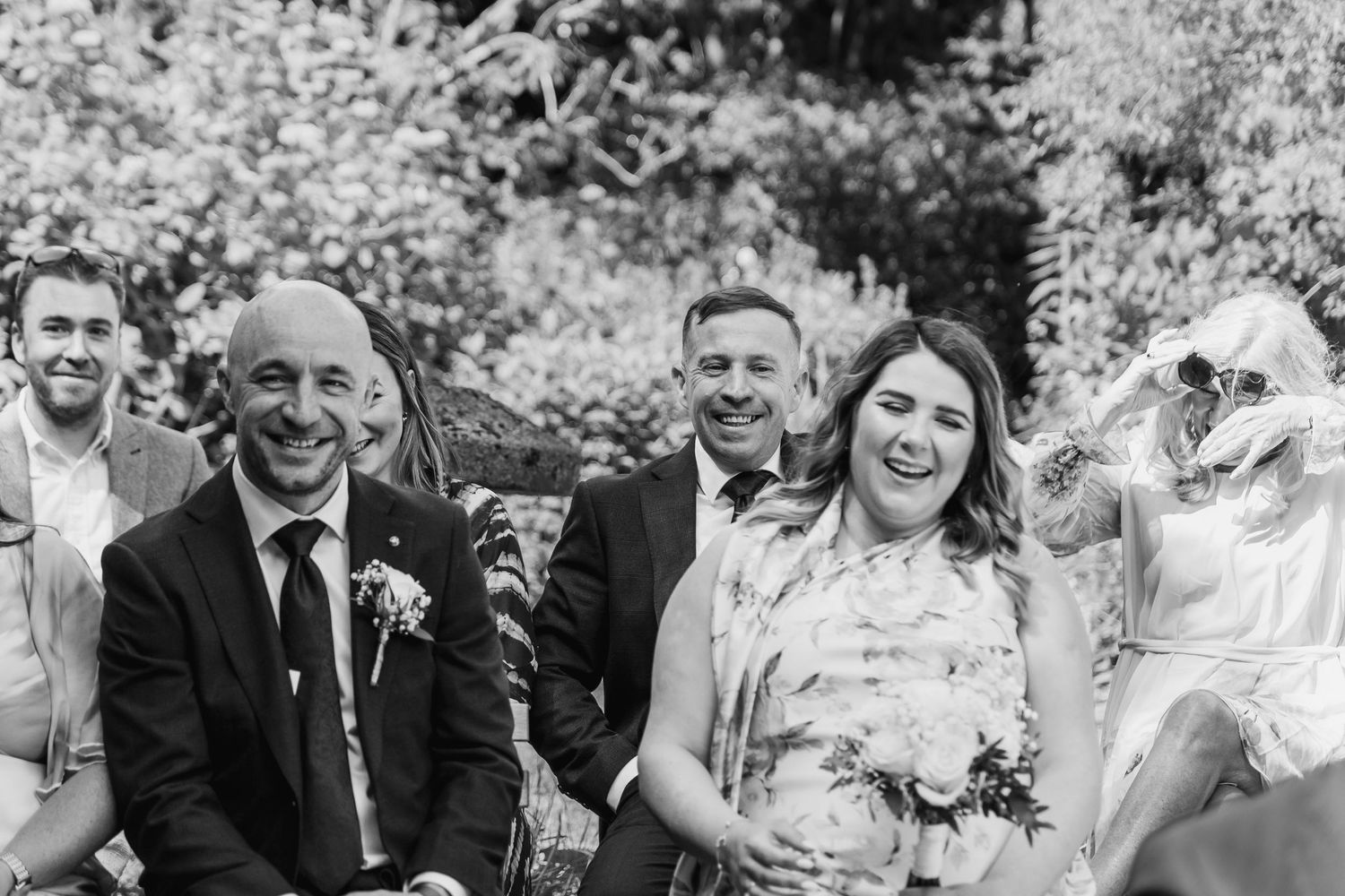 Black and white wedding photo of guests laughing joyfully during an outdoor ceremony.