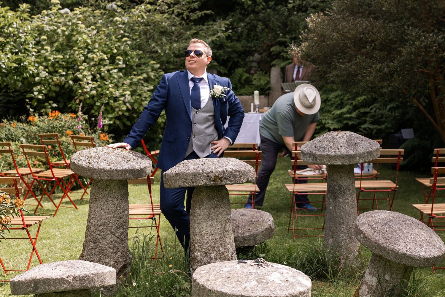 Man in blue suit leaning on stone garden ornament at outdoor wedding ceremony with chairs arranged on lawn.