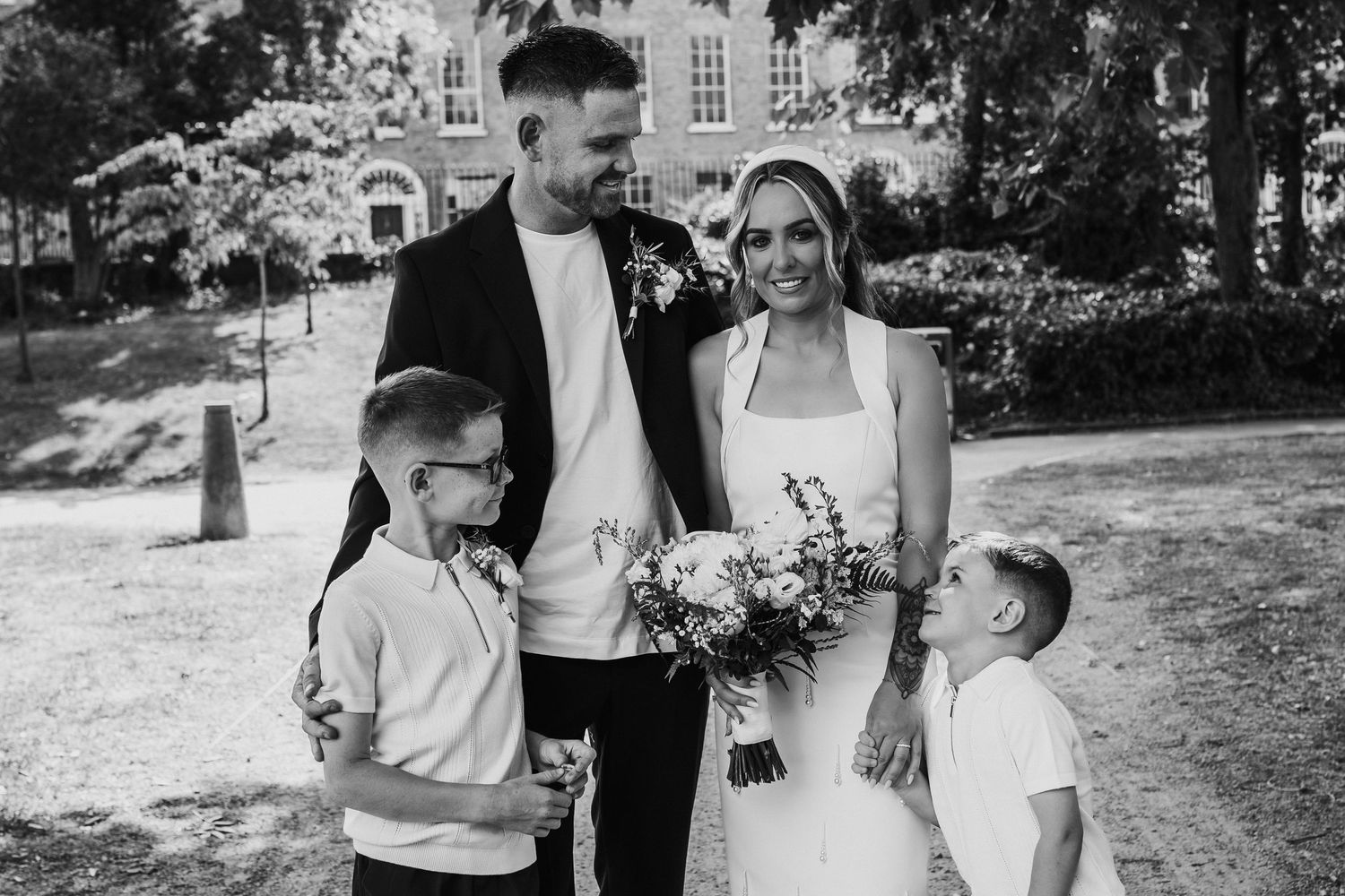 Black and white wedding photo of a couple in formal attire holding a bouquet, posing with two children outdoors.