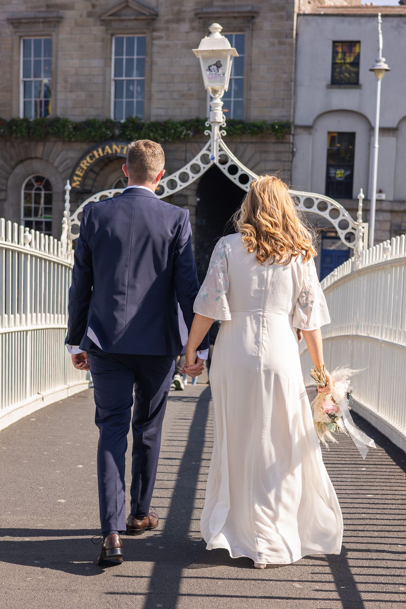 Couple in wedding attire walking together across a historic Dublin city bridge with white railings.