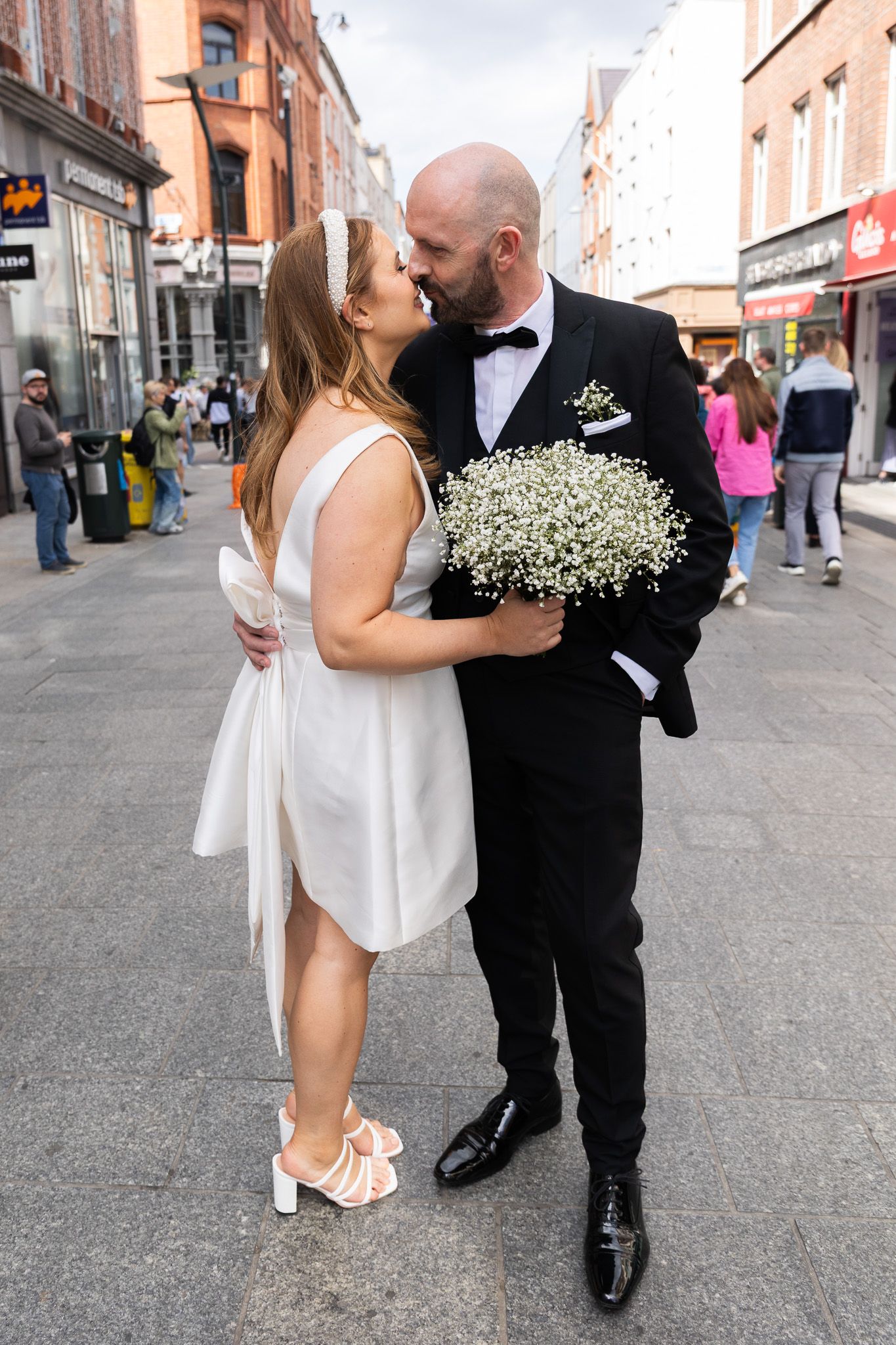 A couple embraces on a busy Dublin city street, with the groom holding a bouquet of baby's breath flowers.