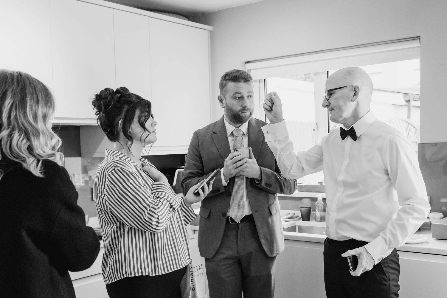 Groom in grey suit gets ready with family in kitchen before wedding ceremony in black and white photo.