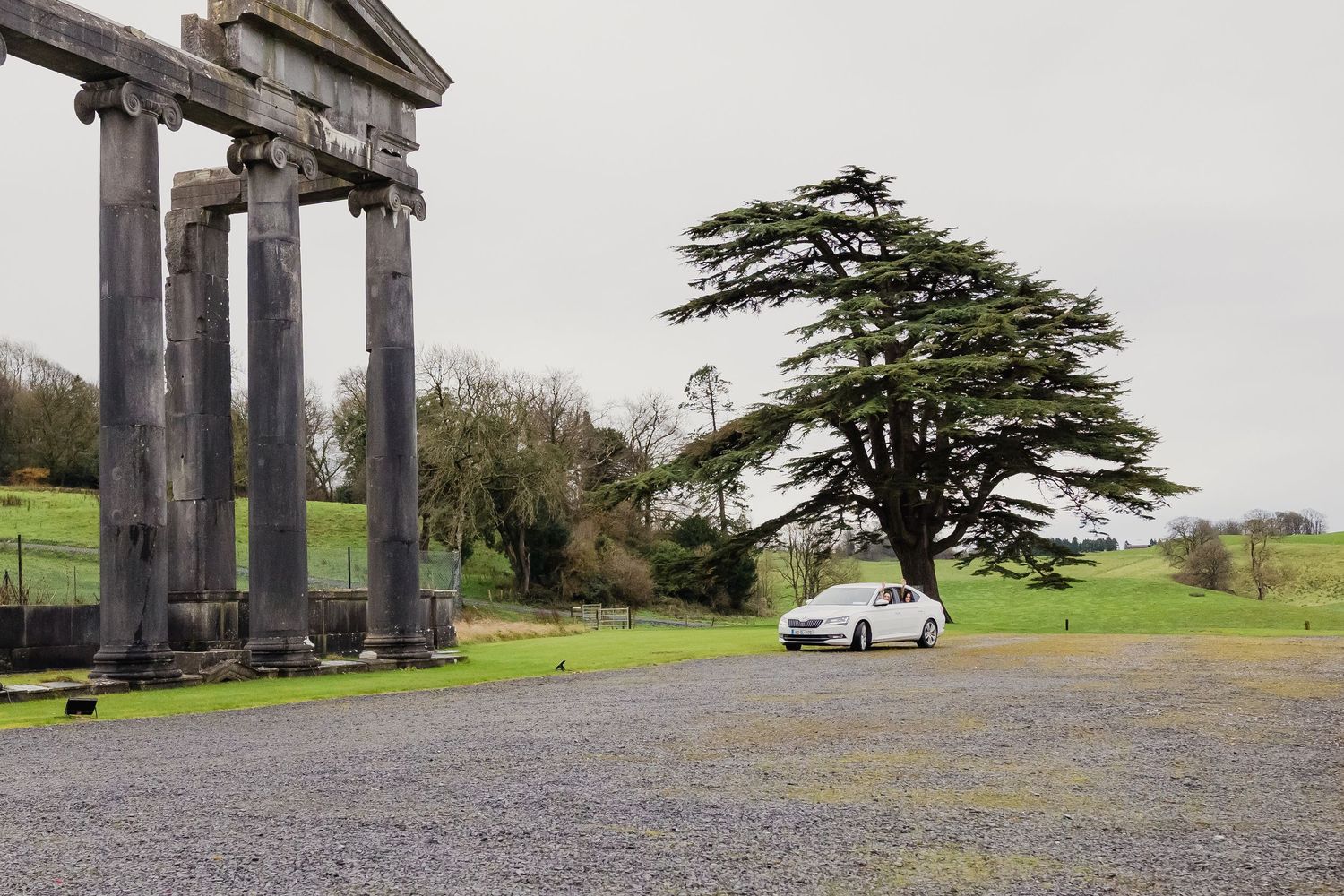 A white car parked near ancient stone columns and a large cedar tree on an overcast day.