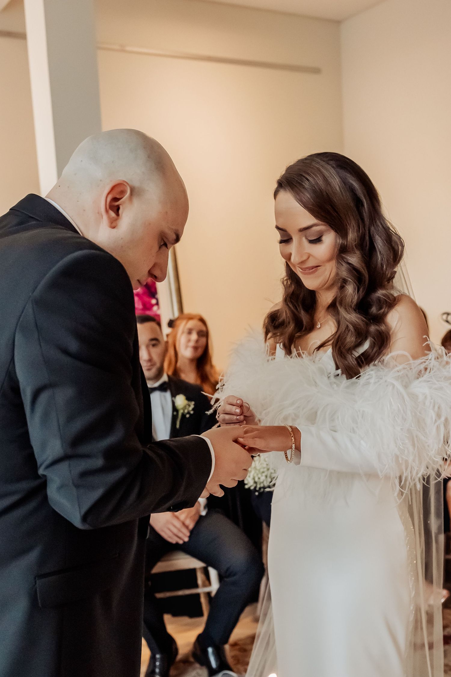 Bride in feather wedding dress exchanges rings with groom in black suit during intimate wedding ceremony.
