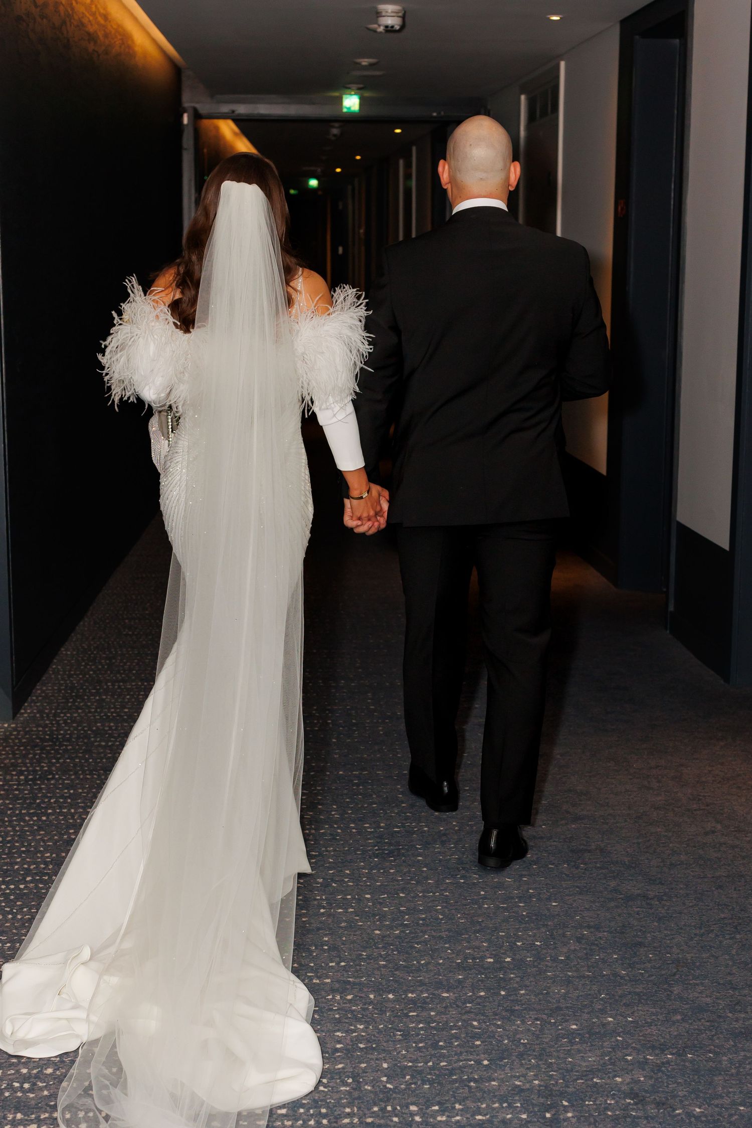 Bride in white feather-trimmed gown and veil walks hand-in-hand with groom in black suit down hotel corridor.