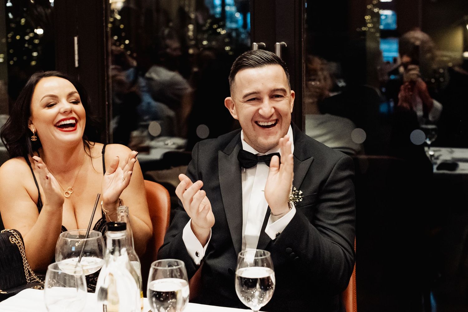 Couple laughing and clapping at a formal dinner event, dressed in elegant black tie attire at a restaurant.