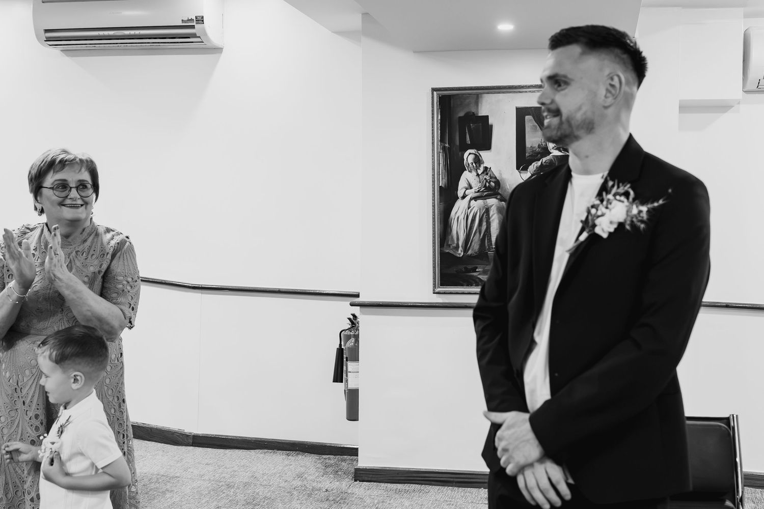 Black and white wedding photo of groom in suit with boutonniere, woman clapping, and child in elegant venue.