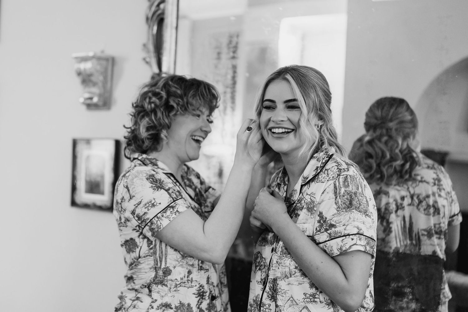 Two bridesmaids in matching floral robes laugh while helping each other with earrings during wedding preparations.