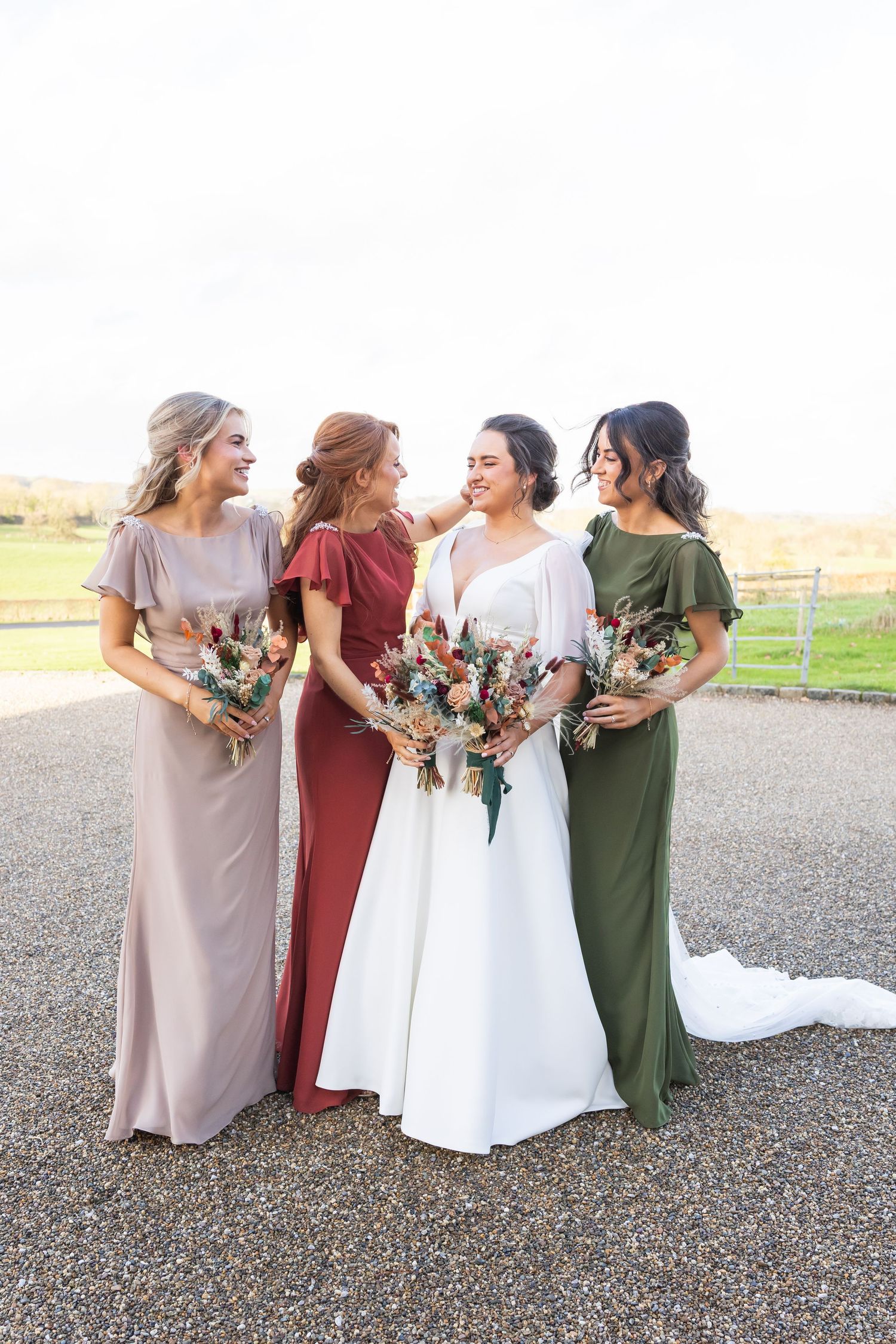Bridesmaids in earth-toned dresses holding bouquets pose together outdoors.