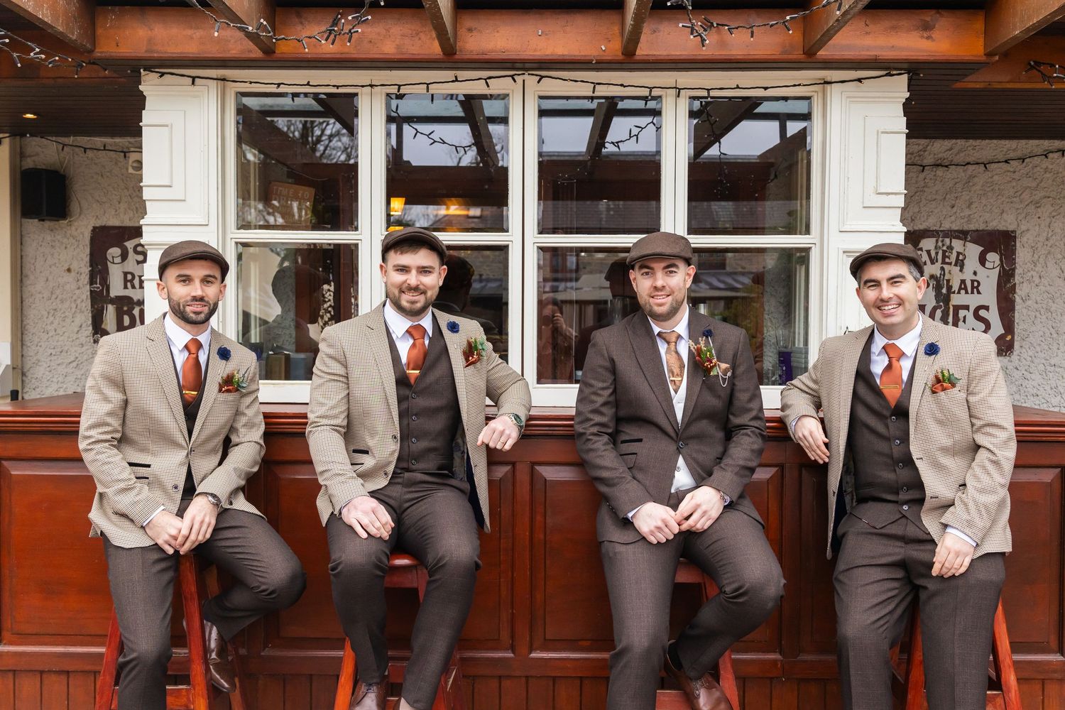 Four groomsmen in matching tweed suits, flat caps, and orange ties pose together outside a rustic pub venue.