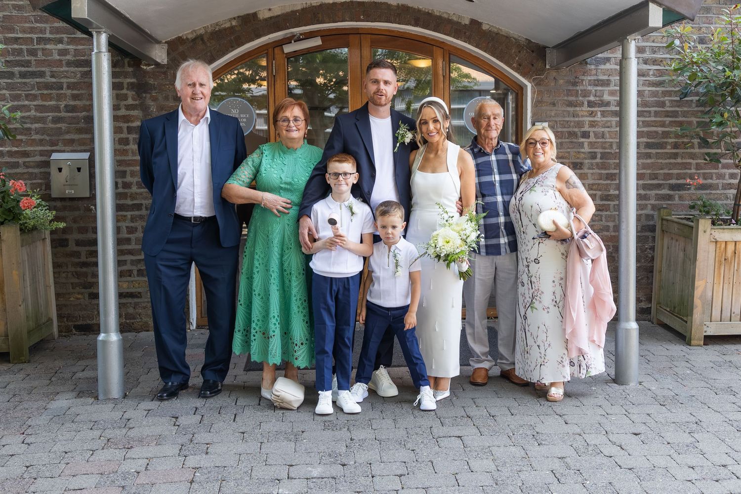 Wedding couple poses with family including two children outside a brick building with wooden doors.