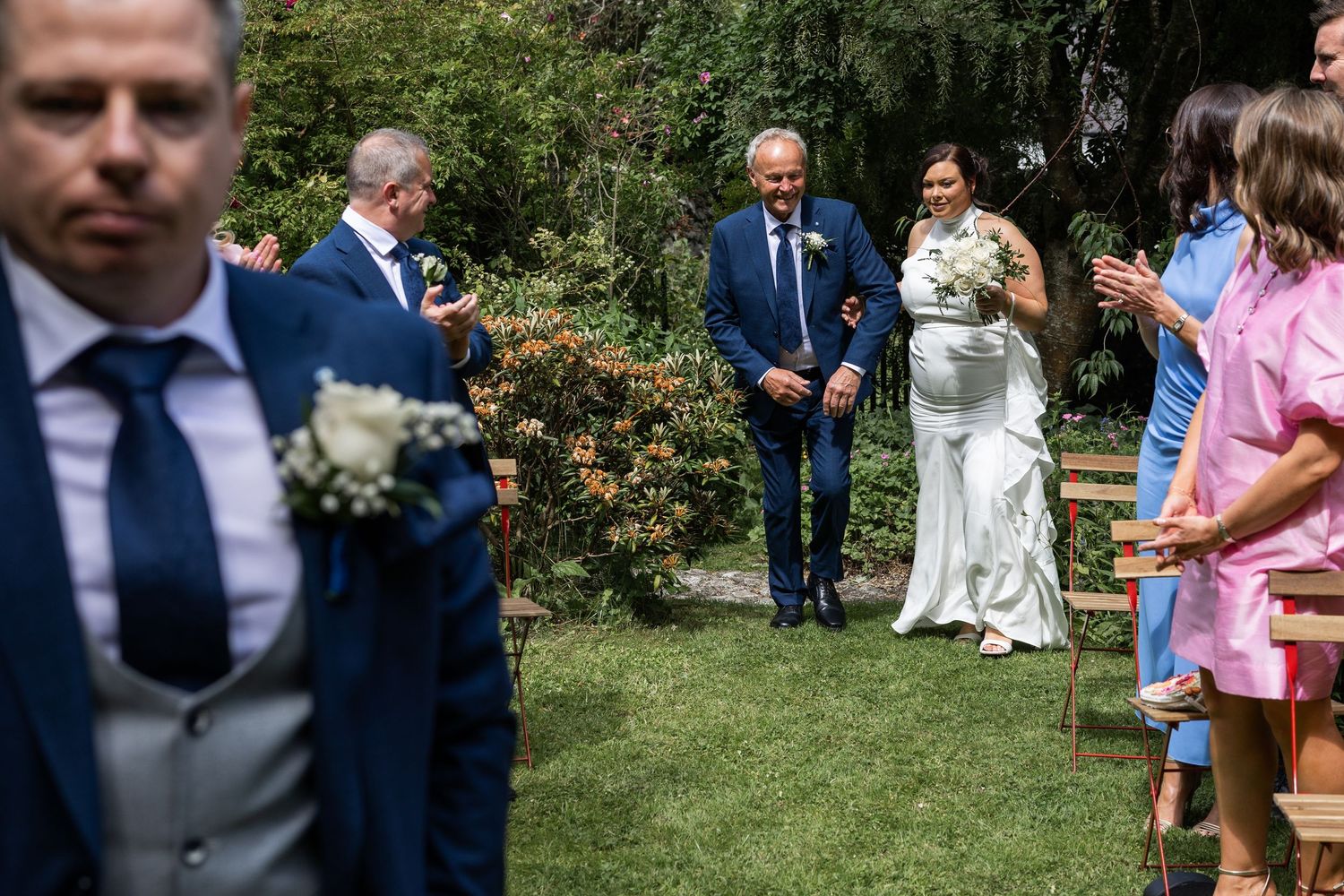Bride walks down outdoor aisle with escort as guests applaud at garden wedding ceremony.